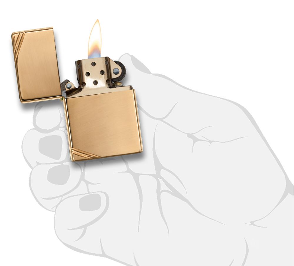 Frontansicht Zippo Feuerzeug Vintage Brass High Polished mit dekorativen Schrägstrichen an beiden Ecken geöffnet mit Flamme in stilisierter Hand